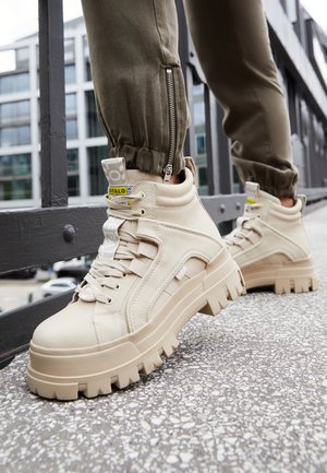 Beżowe wysokie sneakersy na grubej podeszwie z głębokim bieżnikiem, noszone z oliwkowozielonymi joggerami z zamkami, na miejskim chodniku.
