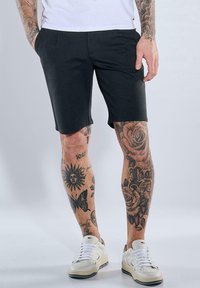 Schwarze Shorts mit maßgeschneiderter Passform, aus glattem Stoff gefertigt, kombiniert mit weißen Turnschuhen. Tattoos zieren die Beine und zeigen florale und Sonnenmotive.