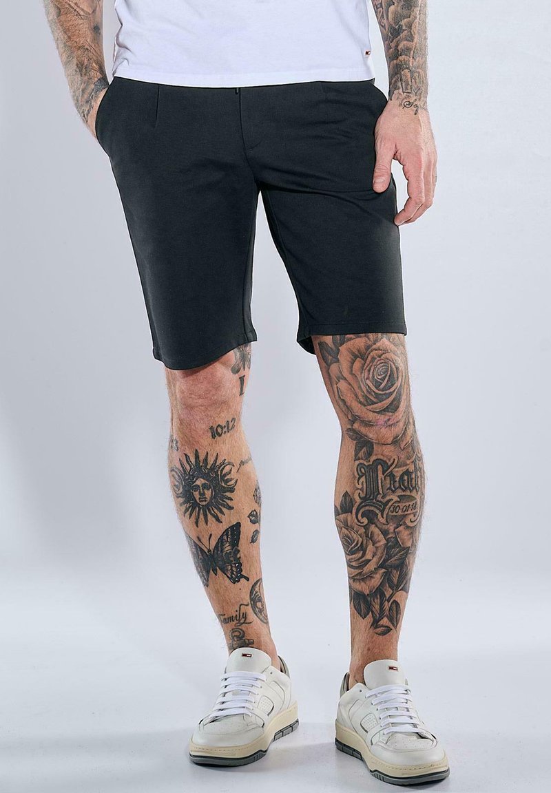 Schwarze Shorts mit maßgeschneiderter Passform, aus glattem Stoff gefertigt, kombiniert mit weißen Turnschuhen. Tattoos zieren die Beine und zeigen florale und Sonnenmotive.
