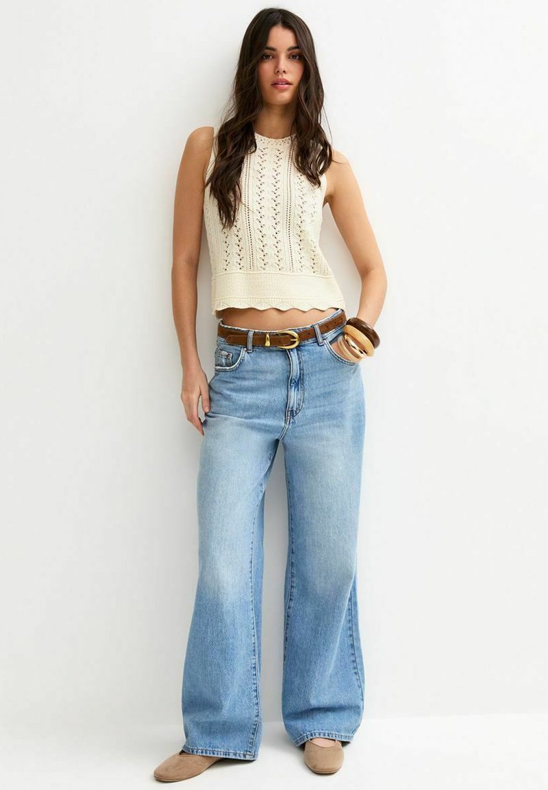 Femme portant un haut en maille crème sans manches, un jean large bleu clair avec une ceinture marron, des chaussures beige et des bracelets en bois sur un fond clair uni.