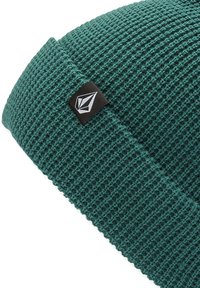 Un bonnet en tricot vert avec un motif texturé. Il présente une étiquette noire avec un logo géométrique blanc sur le côté.