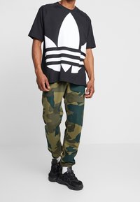 Man med svartvit grafisk t-shirt, grön och brun kamouflagebyxor, vita strumpor och svarta sneakers, som står mot en enfärgad vägg.