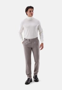 Mann steht mit Händen in den Taschen, trägt einen weißen Rollkragenpullover, beigefarbene Hose und schwarze Anzugschuhe vor einem weißen Hintergrund.