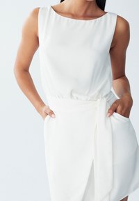 Bílé bezrukávové šaty s kulatým výstřihem, elastickým pasem, detailem na zavázání a bočními kapsami, vyrobené z hladké, tekoucí látky.