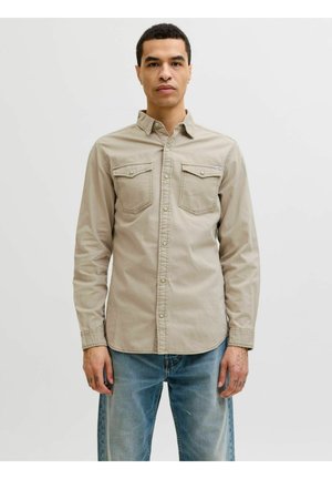 Mann mit kurzen lockigen Haaren trägt beiges Hemd mit Brusttaschen und hellblaue Jeans, steht vor weißem Hintergrund.