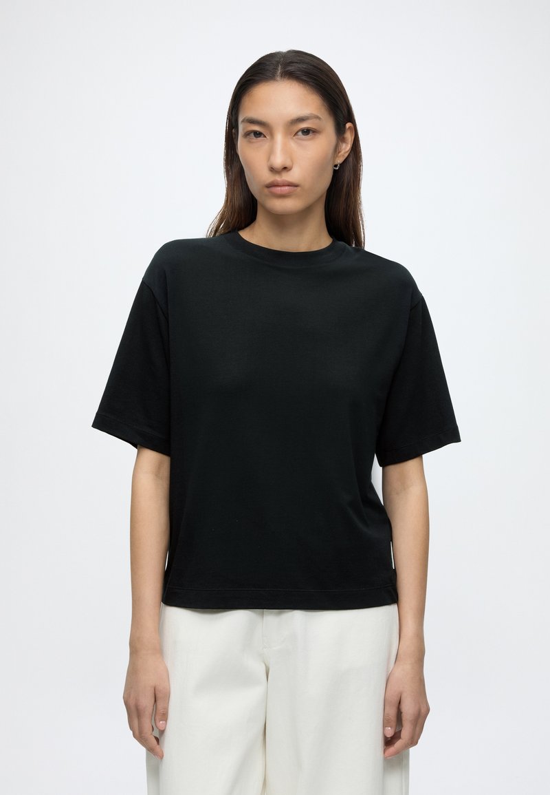 Jeune femme aux cheveux lisses et foncés porte un t-shirt noir uni à manches courtes et un pantalon blanc, debout devant un fond blanc.