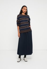 Göndör hajú nő barna és kék csíkos pulóvert, tengerészkék midi szoknyát, szürke zoknit és fehér könnyen felhúzható cipőt visel, miközben egy fehér falnak támaszkodik.