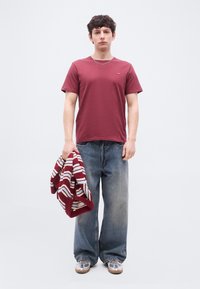 Νεαρός άνδρας με μπορντό μπλουζάκι, φαρδιά μπλε τζιν, ασημένια αθλητικά παπούτσια, κρατώντας μια ριγέ μπορντό και λευκή ζακέτα στο αριστερό χέρι.