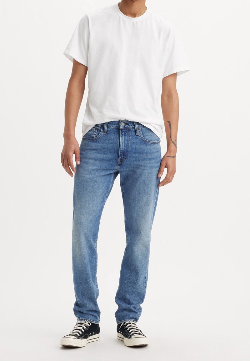 Adulte portant un t-shirt blanc uni, un jean droit bleu clair et des baskets basses noires avec des lacets blancs, debout devant un fond blanc.