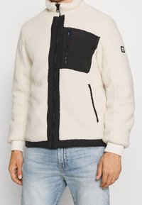 Giacca in pile con corpo crema e pannello nero sul petto, dotata di tasche con zip e polsini a costine. Include chiusura frontale con zip.