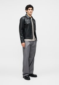 Joven con chaqueta de cuero negra, jersey beige, pantalones anchos grises y zapatos negros, de pie frente a un fondo blanco liso.