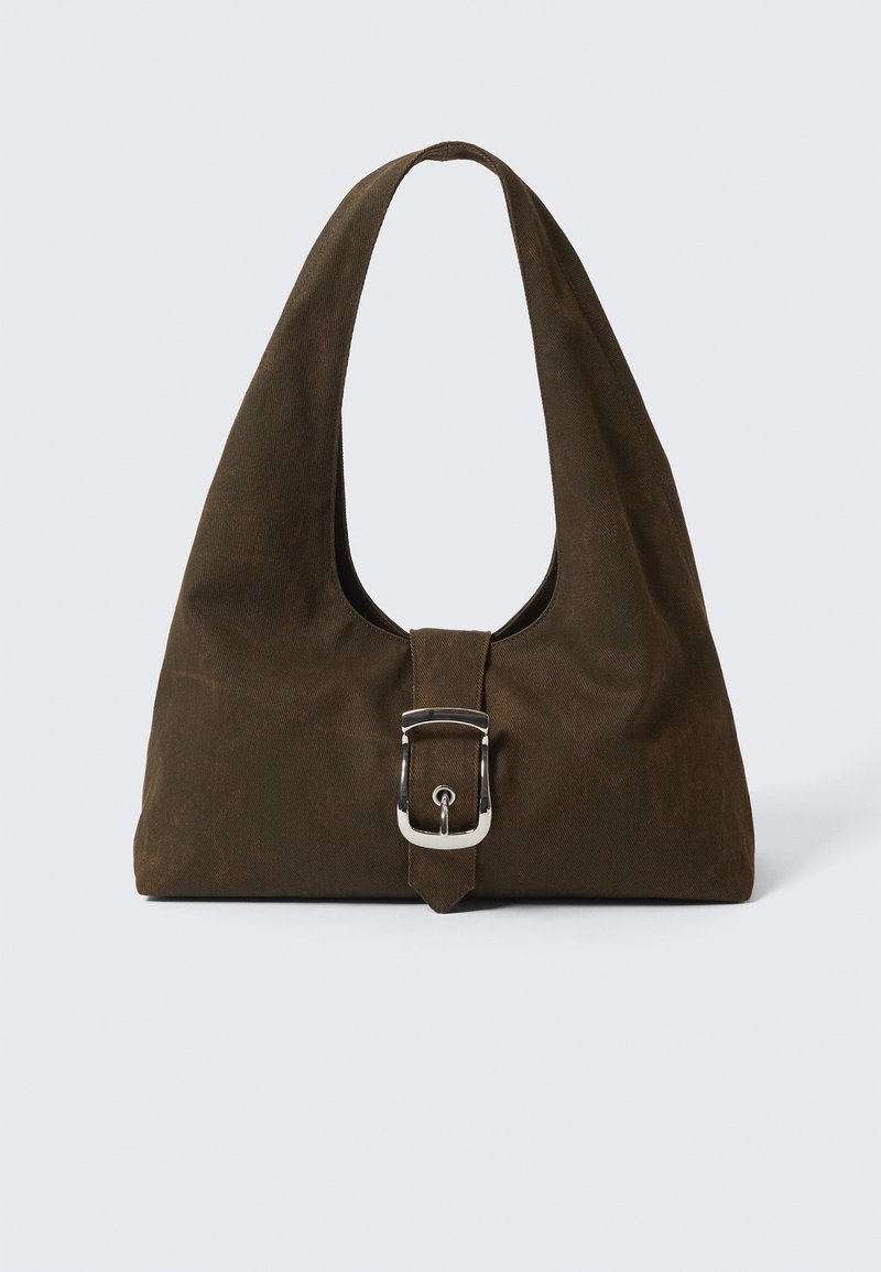 Bolsa de lona marrón con forma de hobo, asa ancha y un acento de hebilla plateada en la parte delantera. Textura suave con un ligero brillo.