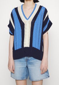 Pull en tricot avec un col en V, featuring des rayures bleues, navy et crème. Les manches courtes et le motif texturé créent une coupe décontractée. Porté avec un short en denim.