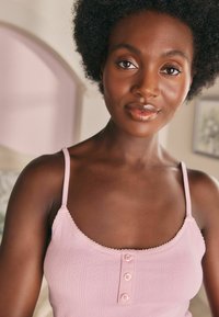 Femme noire aux cheveux crépus naturels portant un haut sans manches rose clair, souriant doucement dans une pièce doucement éclairée au décor neutre.