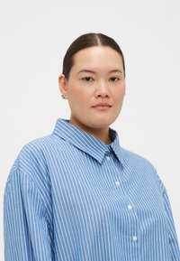 Camisa de botão riscada em azul e branco com colarinho espalhado e mangas longas, apresentando um corte solto e uma textura de tecido macio.