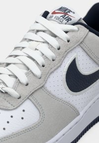 Nike Air Force tenisice s gornjim dijelom od bijele kože i sive suede, perforiranim prednjim dijelom, bijelim vezicama i tamnoplavi akcent logotipa.