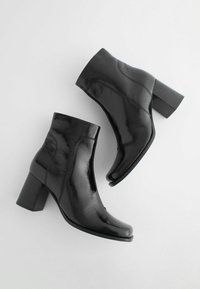 Bottes noires à chevilles en matériau brillant, avec un bout arrondi, un talon bloc et des accents cousus. Texture lisse et design moderne.