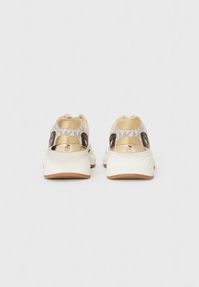 Zapatillas con una parte superior de lona beige que presenta un sutil patrón de logotipos, acentos metálicos dorados y una suela de goma blanca con un bandeado marrón.