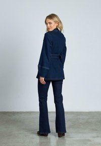 Chaqueta de mezclilla oscura con mangas largas, cintura ceñida con cinturón y collar, combinada con pantalones de mezclilla oscura acampanados. Textura suave, con detalles de costura visibles.