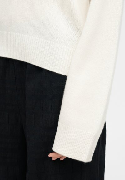 Κοντινό πλάνο ενός ατόμου που φοράει λευκό πλεκτό πουλόβερ με ριμπ μανσέτες και μαύρο παντελόνι με υφή, με το χέρι χαλαρό στο πλάι.