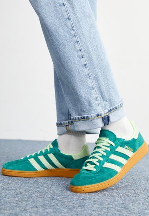 Persoon draagt teal groene Adidas sneakers met rubberen zolen en witte strepen, lichtblauwe opgerolde jeans en witte sokken, staand op tapijt.