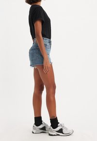 Black short-sleeve t-shirt, denim shorts with frayed edges, side view. Silver athletic shoes with black accents and black socks.