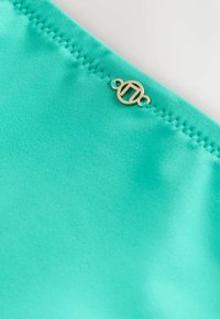 Tissu de maillot de bain turquoise à texture lisse, avec un accent doré et un logo circulaire près de la couture.