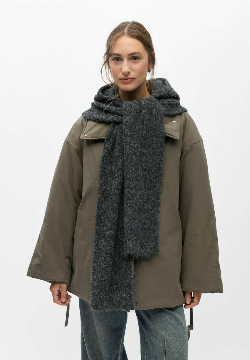 Jeune femme portant une veste taupe oversize, une grande écharpe gris foncé nouée autour du cou, avec un jean bleu, debout sur un fond blanc uni.