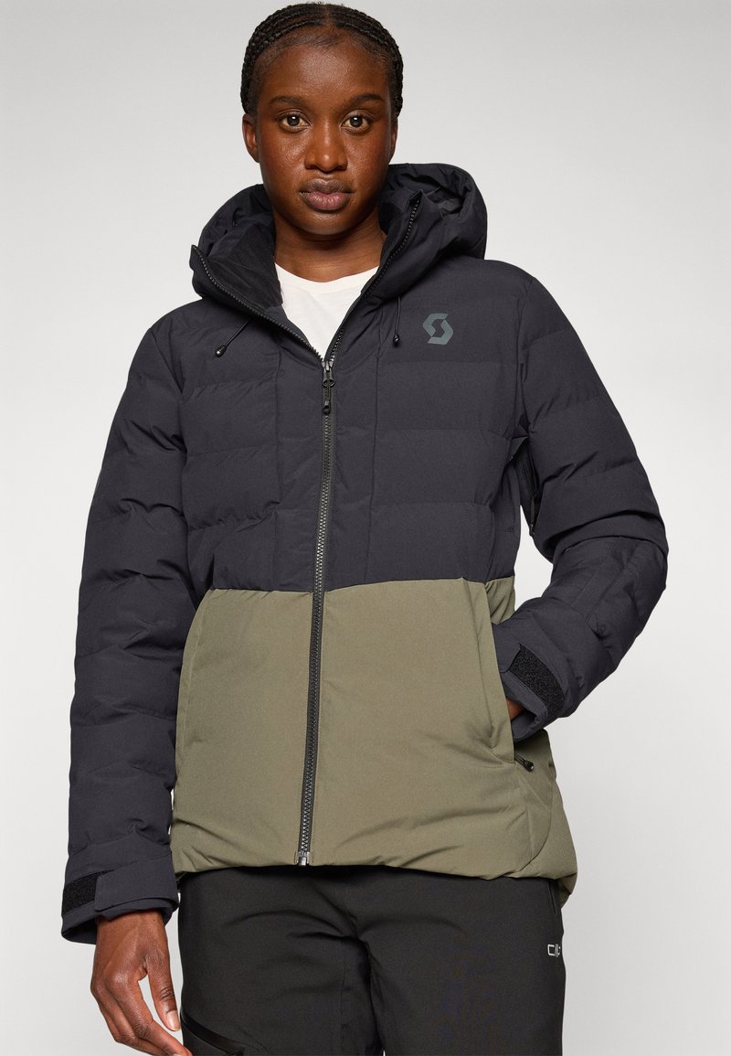 Person, die eine zweifarbige schwarze und olivgrüne Steppjacke mit Kapuze trägt und mit einer Hand in der Tasche an einem schlichten Hintergrund steht.