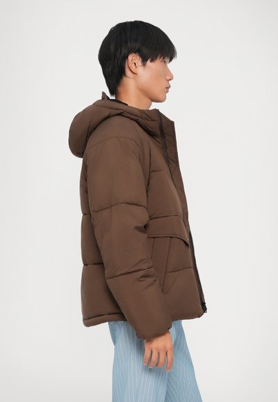Giacca marrone imbottita con cappuccio, dotata di chiusura a zip e tasche frontali. Il tessuto è liscio con una texture trapuntata, indossata sopra pantaloni a righe chiare.