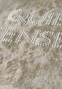 Béžová látka s vyvýšenými stříbrnými hřebíky tvořícími text, s texturovaným pozadím a jemnými barevnými variacemi. Slova zahrnují "TIŠE" a "VĚDOMÍ."