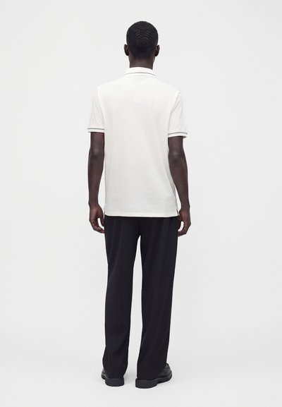 Homme debout, dos à la caméra, portant un polo blanc à manches courtes, un pantalon noir et des chaussures noires sur un fond clair uni.