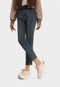 Sneaker beige con tessuto testurizzato e dettagli bianchi, indossati con jeans in denim scuro con vestibilità affusolata e minima sbiaditura.