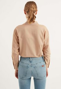 Camicia button-up beige chiaro con maniche lunghe, abbinata a jeans azzurri a vita alta con tasche posteriori e un accento in pelle con logo.