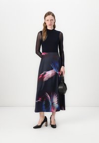 Haut noir à manches longues avec des bras transparents, associé à une jupe fluide bleu marine ornée d'un imprimé coloré de plumes. Talons noirs et petit sac.