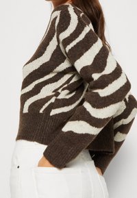 Woman wearing a brown and cream zebra-patterned sweater tucked into white pants, hands resting in back pockets, side profile visible.