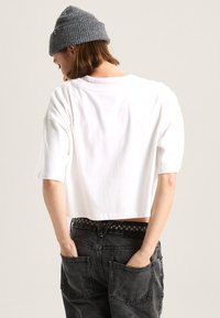 Camiseta blanca recortada de manga corta, acompañada de unos jeans gris oscuro con grandes bolsillos y un cinturón negro con patrón de cuadros.