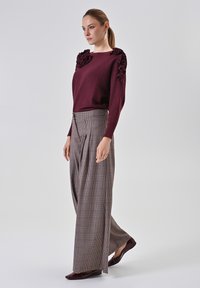 Weinrote Sweatshirt mit gerüschten Schulterakzenten, kombiniert mit hoch taillierten beigen Karohosen und weinroten Ballerinas. Durchgehend glatte Texturen.