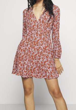 Femme portant une robe fleurie couleur rouille avec des manches longues et un décolleté en V, debout devant un fond clair uni.