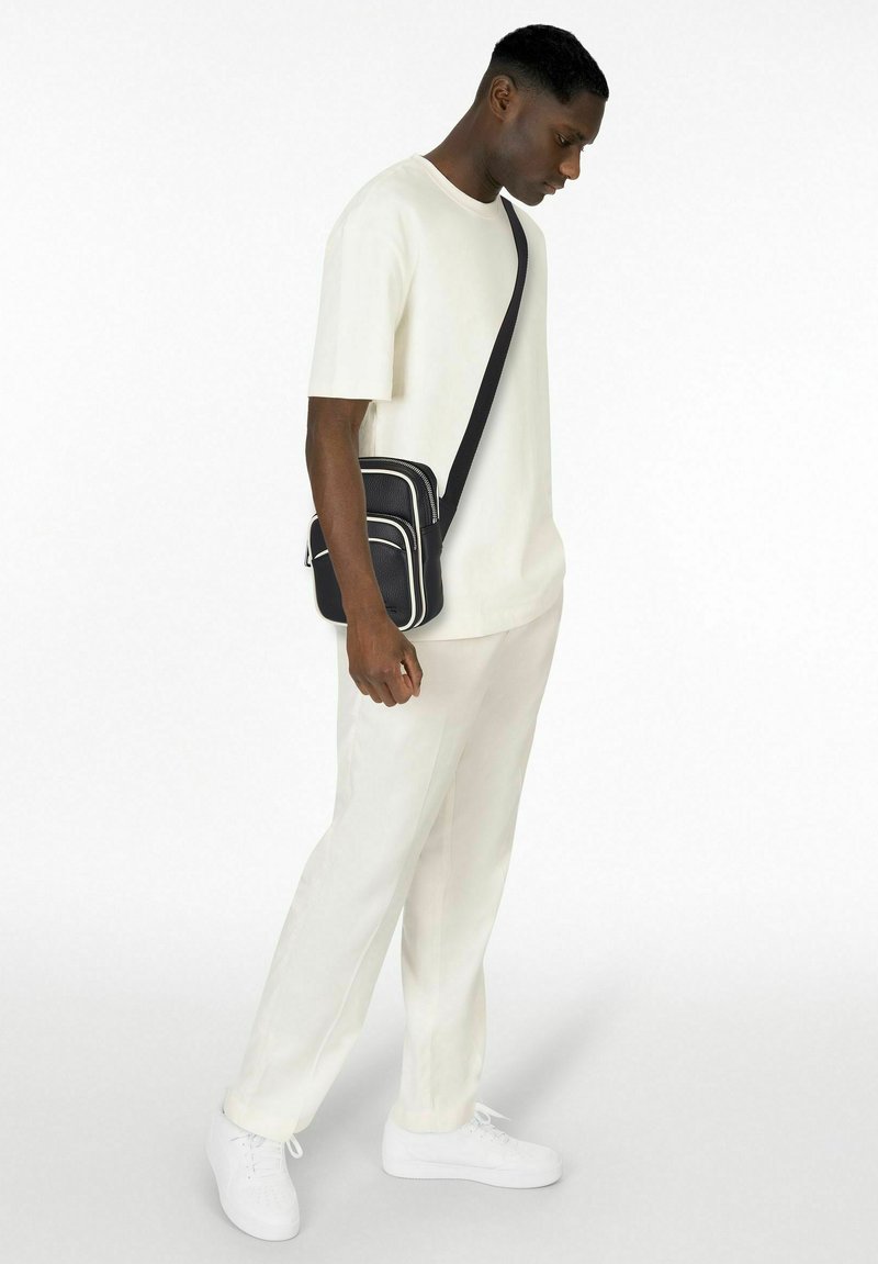 T-shirt blanc à manches courtes, coupe décontractée, associé à un pantalon de couleur claire. Un sac bandoulière noir avec des bordures blanches et une surface texturée.