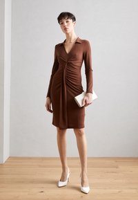 Robe longue marron à manches longues avec un décolleté en V et un design froncé, assortie à des chaussures à talons blancs et à une petite pochette blanche.