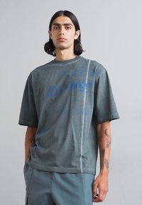 T-shirt oversize gris à manches courtes, avec un imprimé graphique bleu et un détail de couture contrasté sur un côté. Tissu en coton doux.