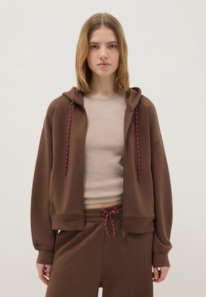 Junge Frau trägt braunen Kapuzenpullover mit Reißverschluss und passende Jogginghose mit burgunderroten Kordeln, beige geripptes Top, steht vor schlichtem Hintergrund.