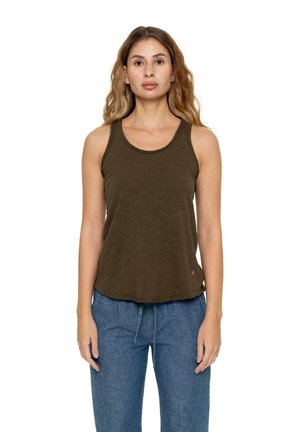 Une femme porte un haut sans manches vert olive foncé et un jean bleu sur un fond blanc uni.