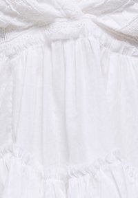 Tissu blanc avec des parties froncées et volantées présentant une délicate bordure en dentelle et des motifs brodés à jours.