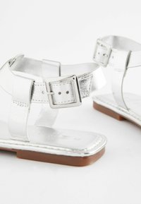 Silver metallic sandaler med justerbara ankelremmar, platta bruna sulor och en slät yta, med spänne och minimalistisk design.