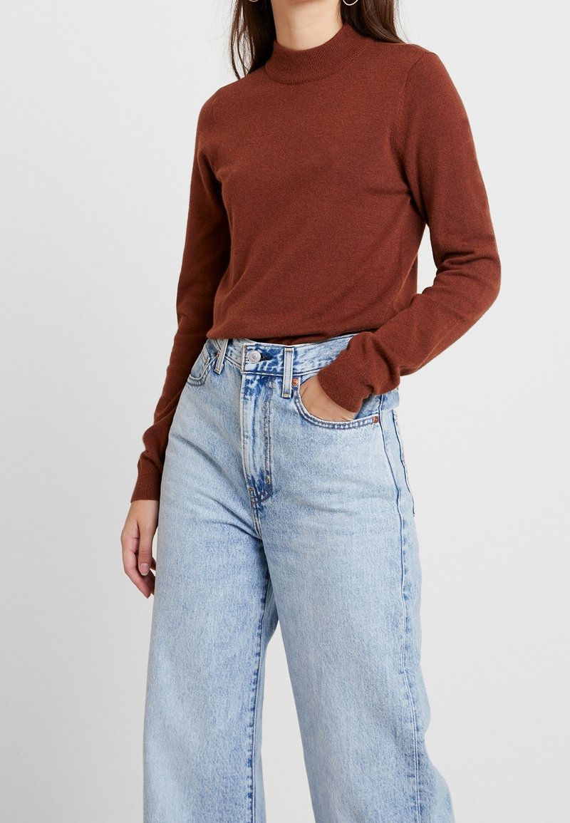 Femme portant un pull à col roulé couleur rouille, rentré dans un jean taille haute à jambes larges bleu clair, avec la main dans la poche.