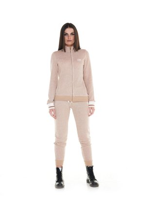 Une femme porte un survêtement beige composé d’une veste zippée et d’un pantalon de jogging assorti, ainsi que des bottes noires à lacets, sur fond blanc.