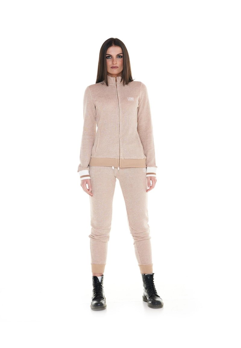 Une femme porte un survêtement beige composé d’une veste zippée et d’un pantalon de jogging assorti, ainsi que des bottes noires à lacets, sur fond blanc.