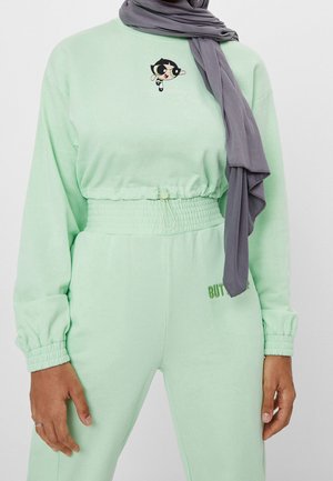 Person trägt minzgrüne Jogginghose mit elastischem Bund und passendes langärmeliges Oberteil mit kleinem Powerpuff Girls-Logo auf der Brust, grauer Hijab.